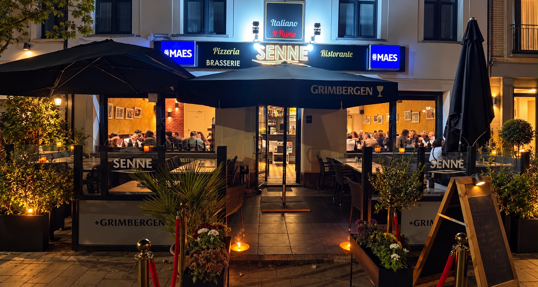 Terrasse illuminée du restaurant Il Fiume Senne vue de face le soir, parasols et enseigne SENNE